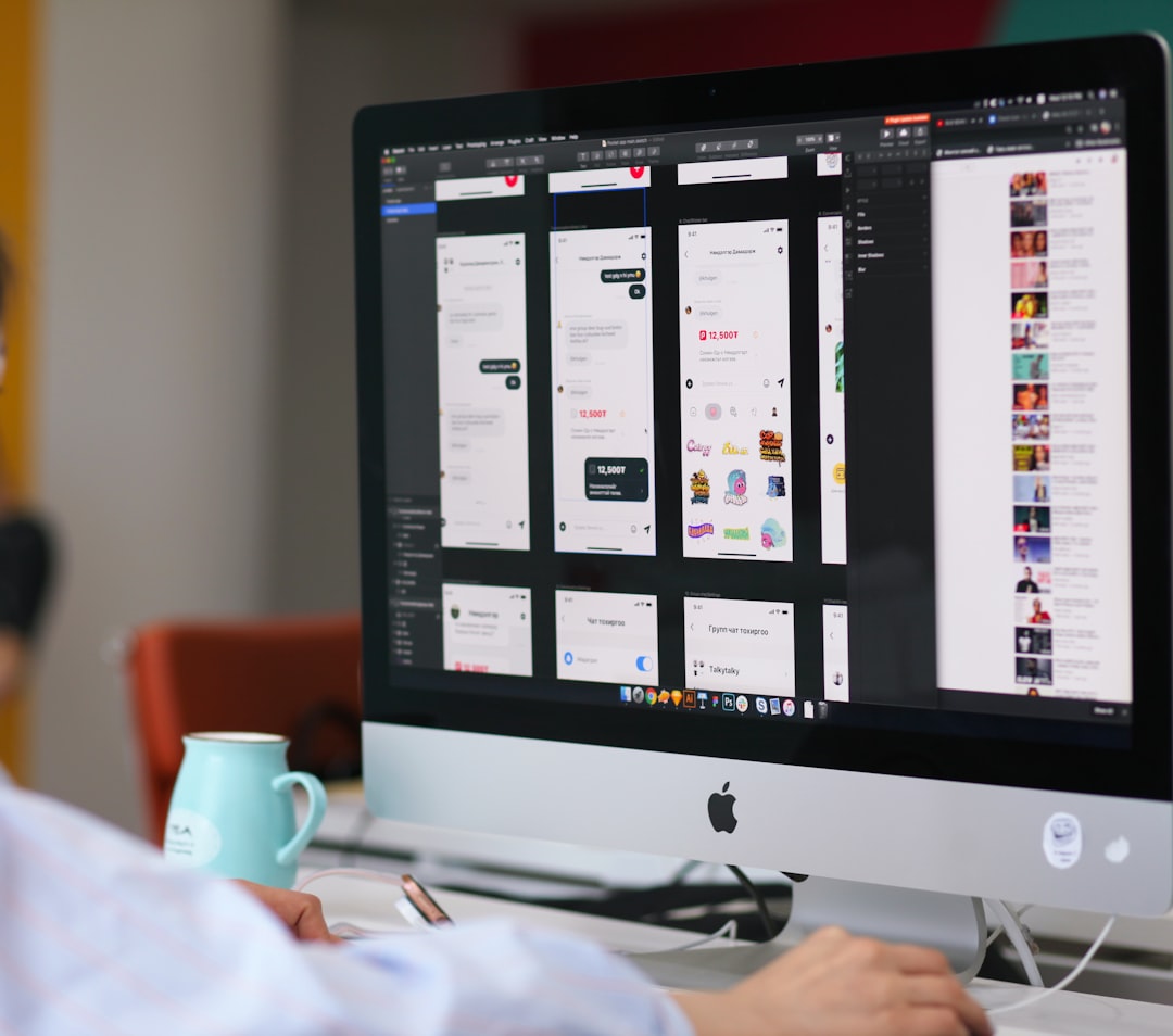 Person working at an iMac, editing mobile app UI screens in a design tool (wireframes and mocks) on a desk with a mug nearby.