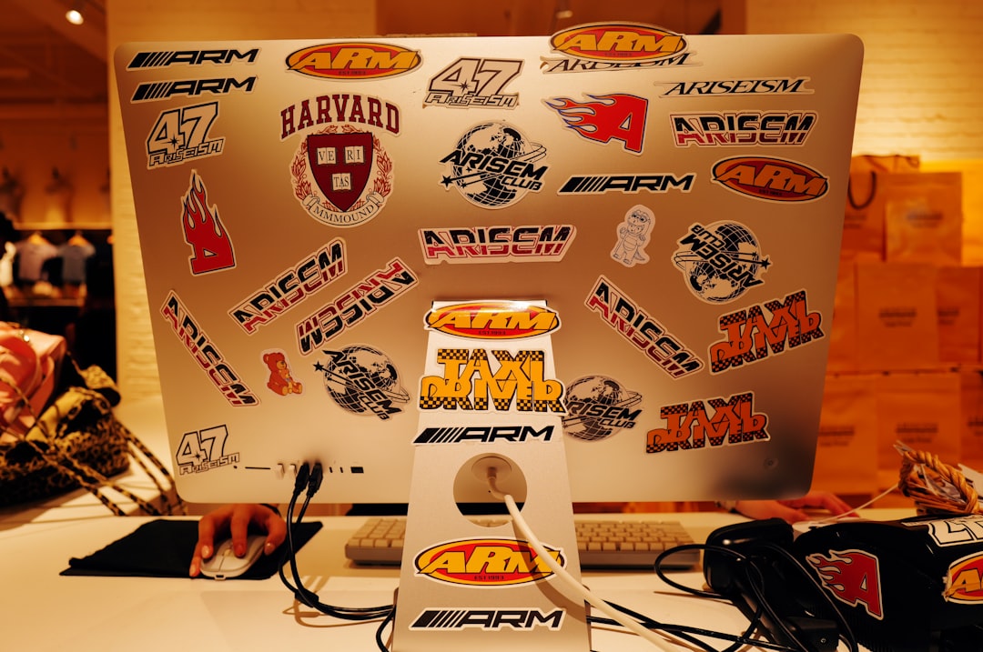 Laptop monitor covered in stickers for ARM, ARISEM, Harvard, and related brands on a desk with keyboard and mouse nearby.