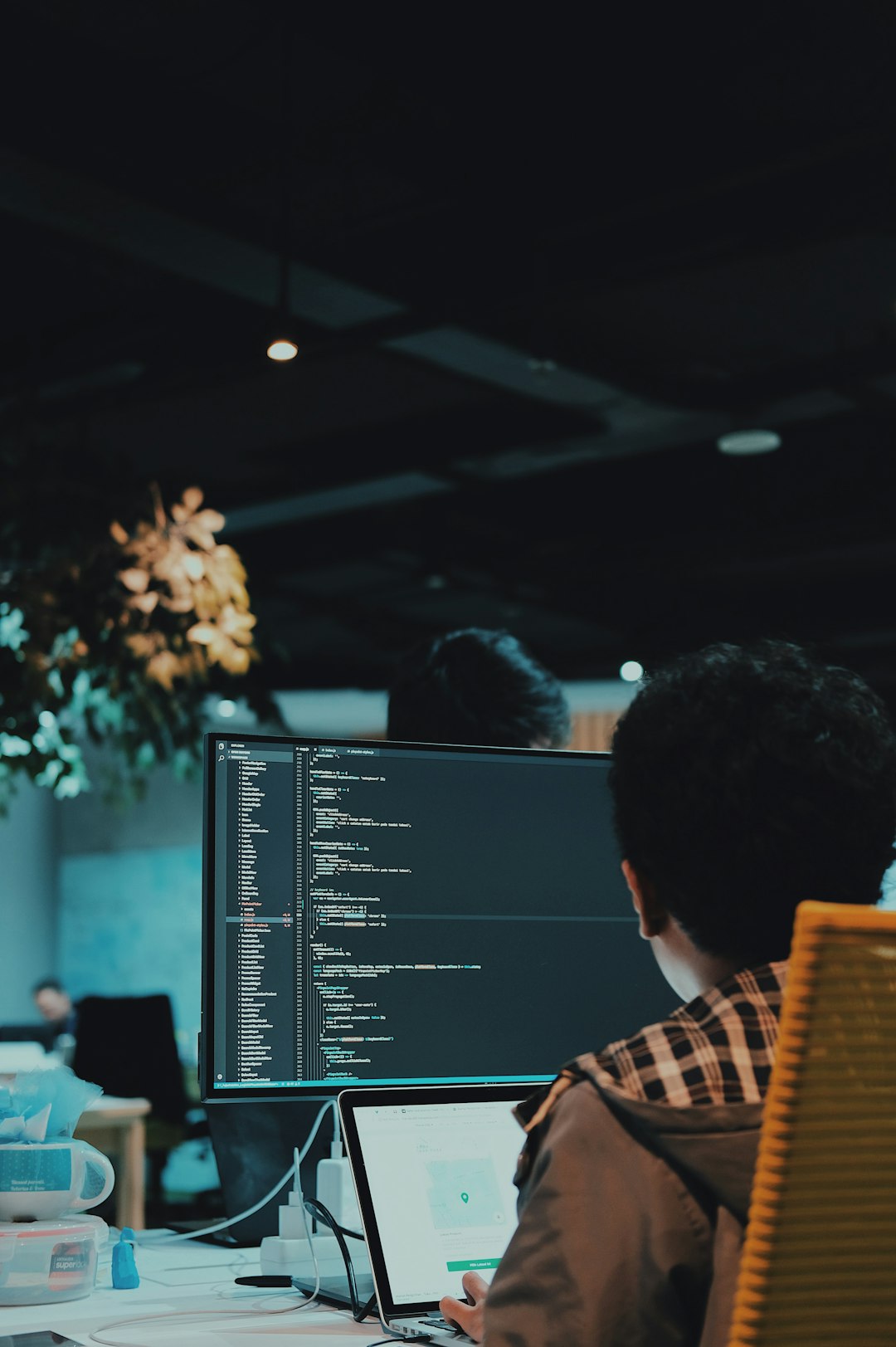 Coder at a workstation reviewing lines of code on a large monitor in a dim office environment, laptop open nearby and office plants in the background