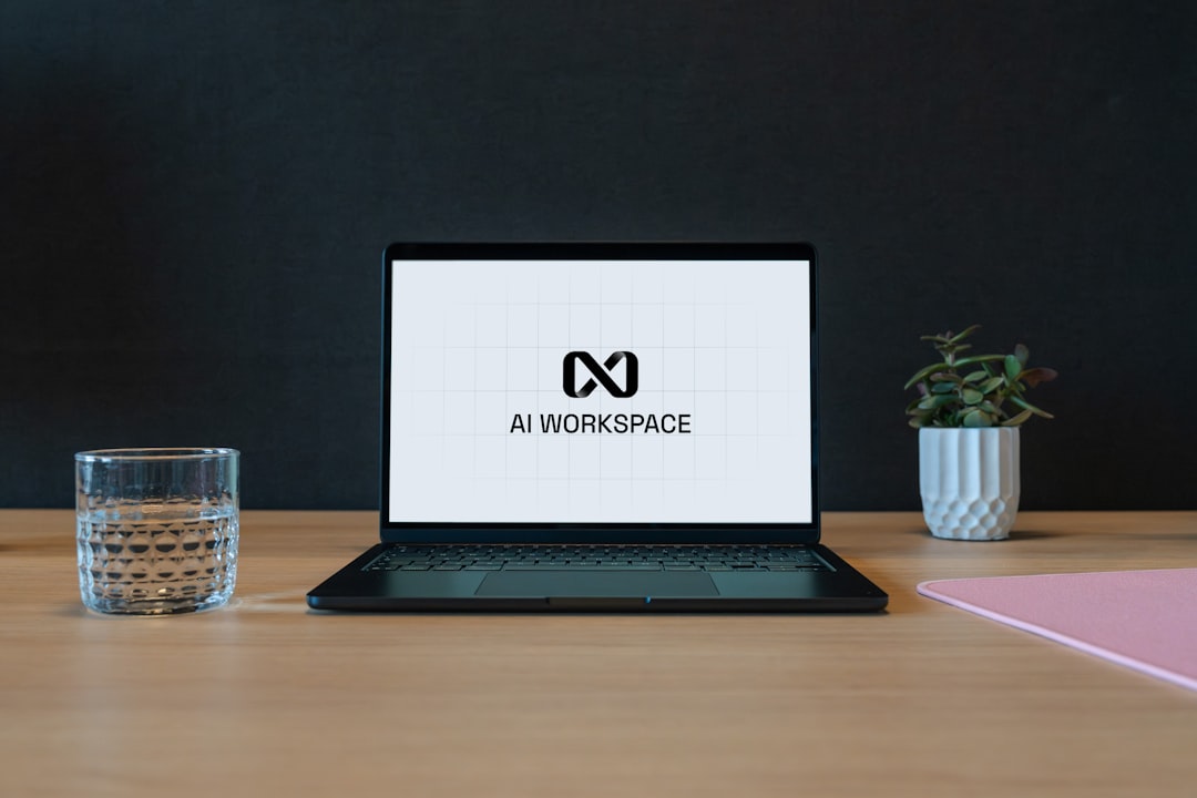 Laptop on a wooden desk displaying the AI WORKSPACE logo on screen, with a glass, small plant, and pink notebook nearby.