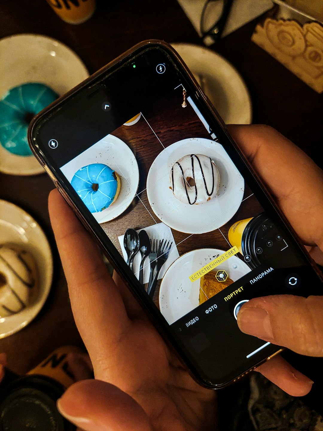 Hands hold a smartphone taking a photo of two donuts on white plates (blue glazed donut and white with chocolate drizzle) at a table, with a yellow label 'ЕСТЕСТВЕННЫЙ СВЕТ' on the screen.