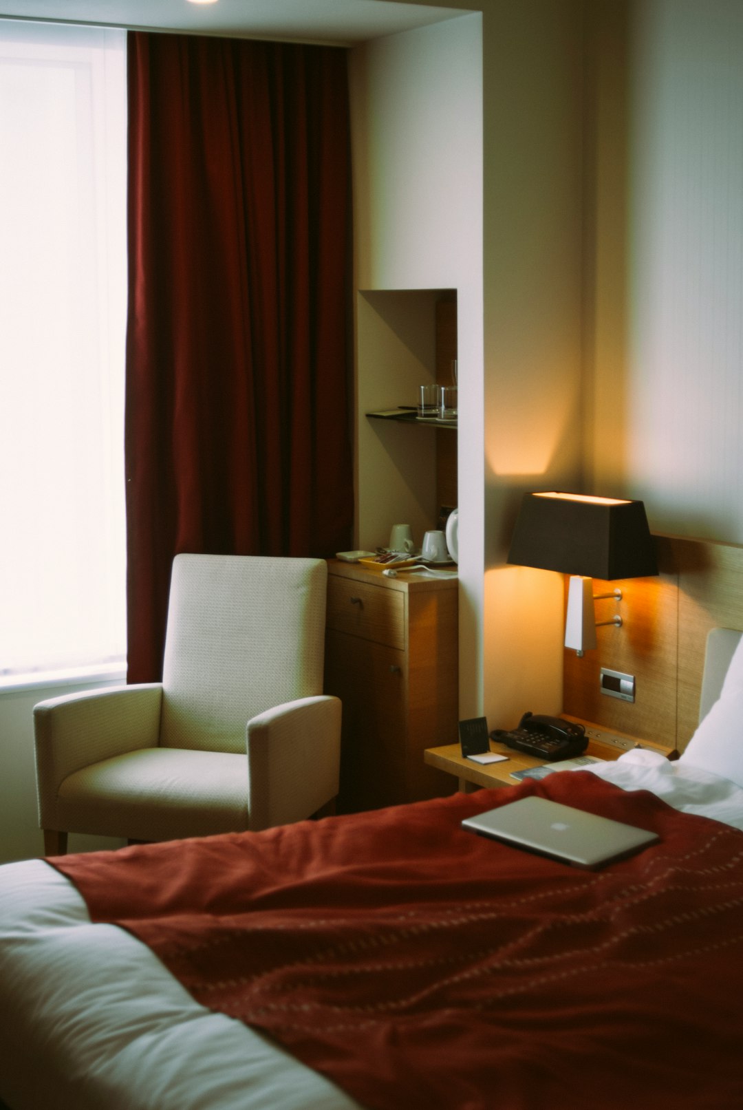 Hotel room corner: bed with a red duvet in the foreground, beige armchair by a window with red curtains, and a wooden desk nearby.