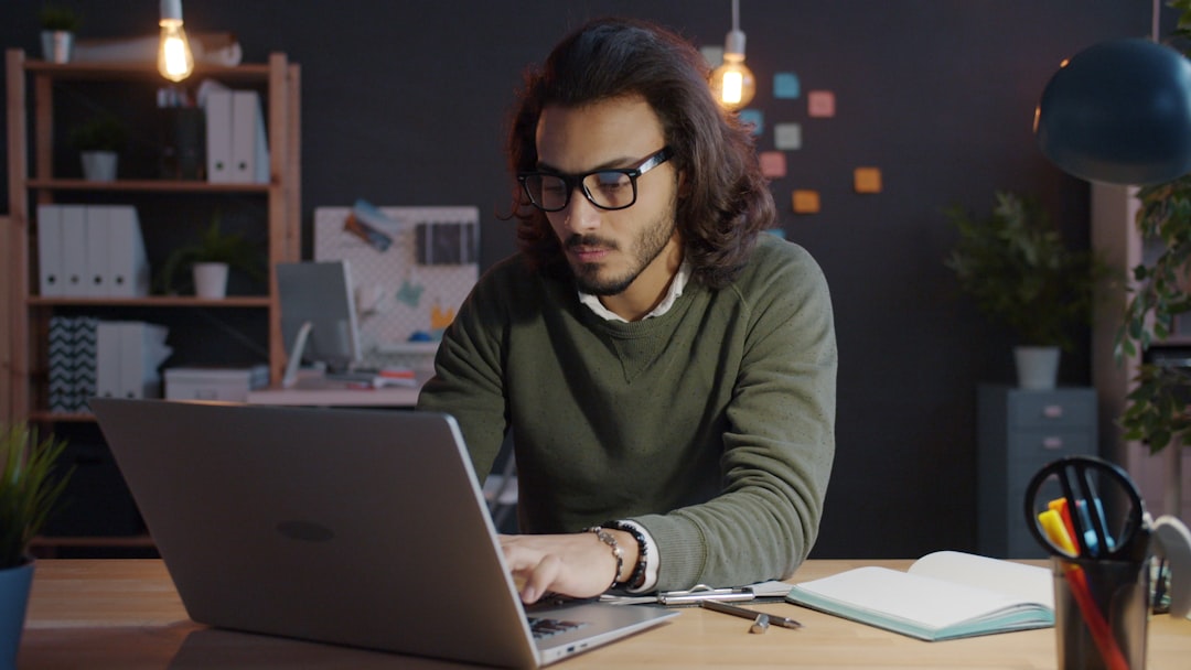 Man with glasses typing on a laptop at a desk in a modern office, notebook and pens nearby.