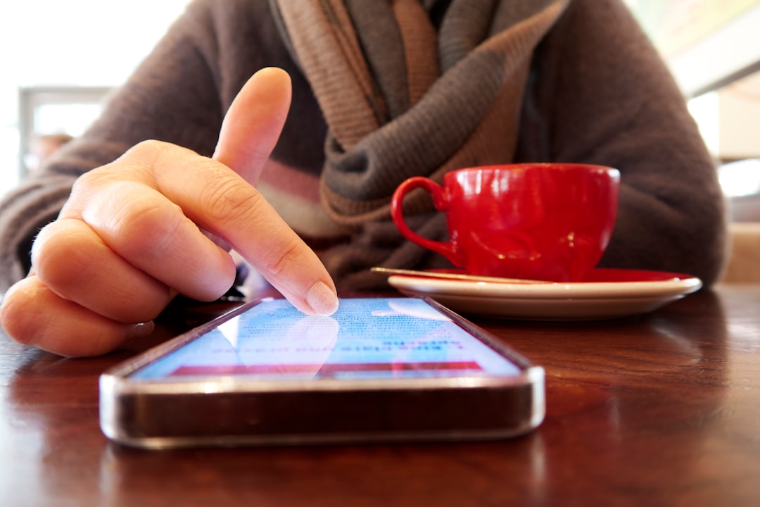 Person in a brown sweater taps a smartphone on a wooden cafe table, with a red coffee cup on a saucer nearby.” Note: remove this stray bracket.