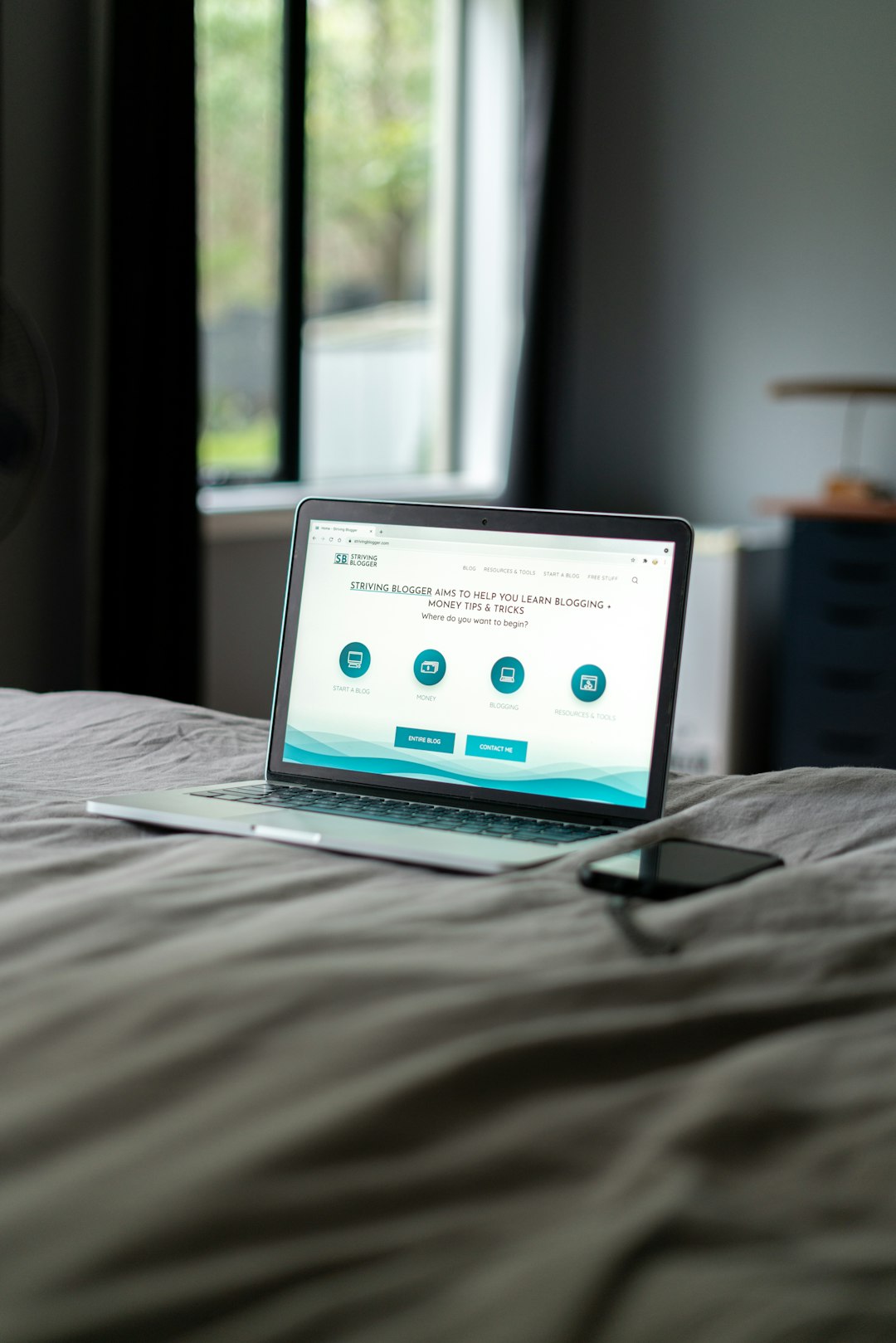 Laptop on a bed open to a blog homepage with blue circular icons, a smartphone beside it.