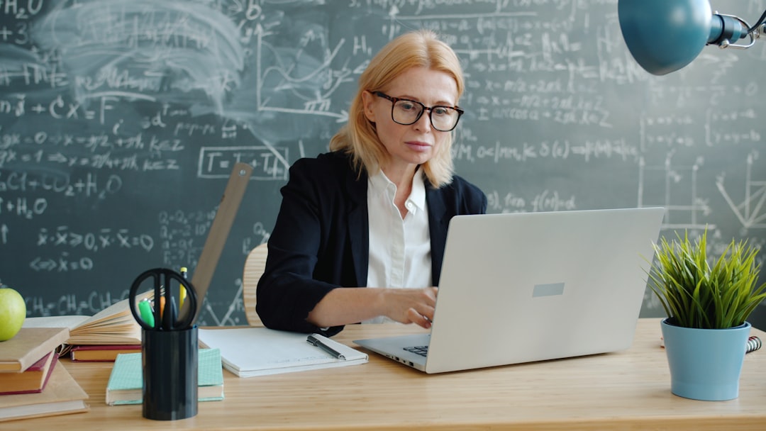 Woman in glasses works on a laptop at a classroom desk, chalkboard filled with equations behind her.