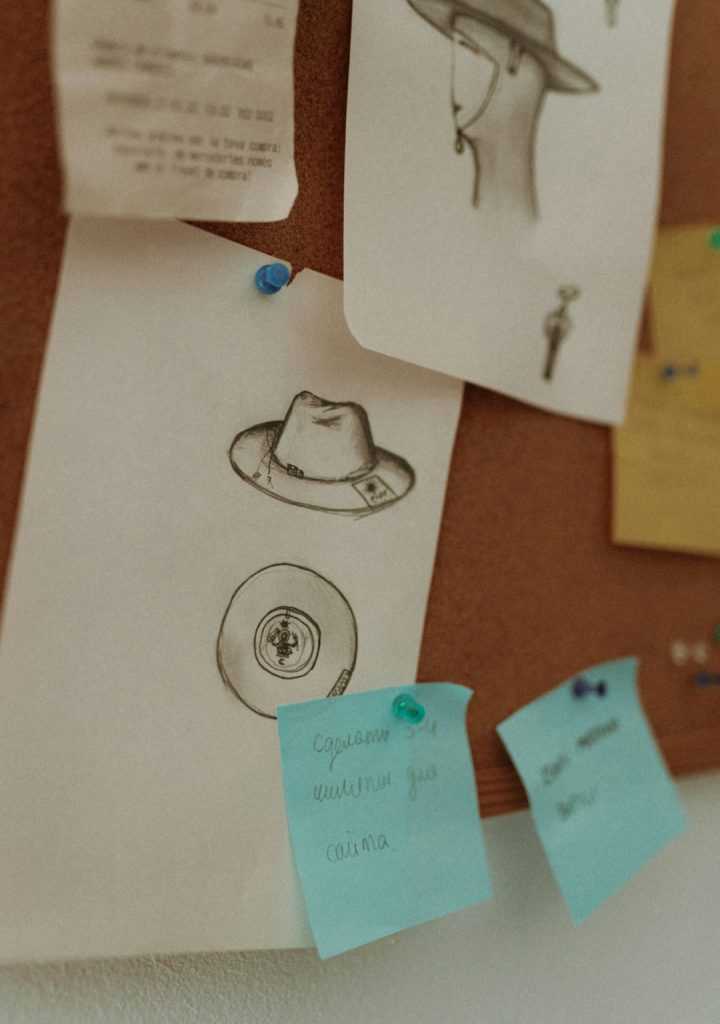 Mood board with hand-drawn hat and badge sketches pinned to a corkboard, blue sticky notes with handwritten notes nearby.