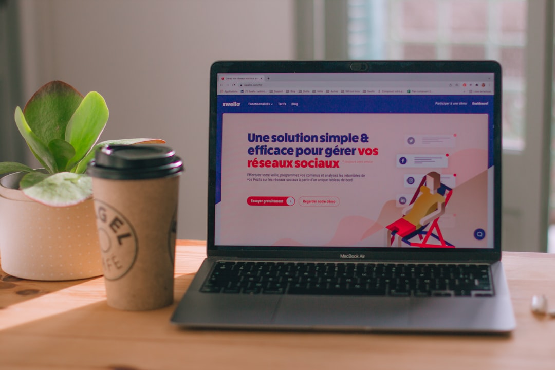 Laptop on a wooden desk showing a French social media management site, with a potted plant and a takeaway coffee cup beside it.