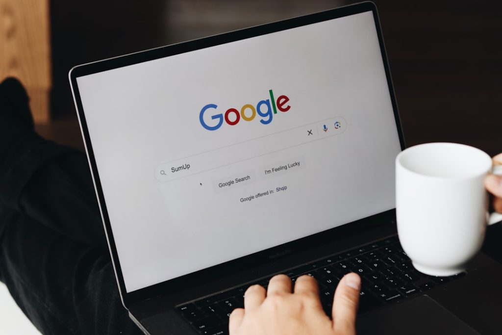 Person using a laptop with the Google homepage visible on screen and a white mug resting beside the keyboard on the right.