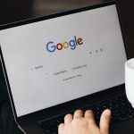 Person using a laptop with the Google homepage visible on screen and a white mug resting beside the keyboard on the right.