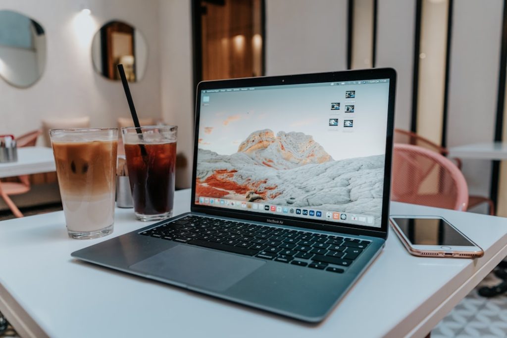 Laptop open on a white table with two iced coffees and a pink smartphone in a bright cafe setting.