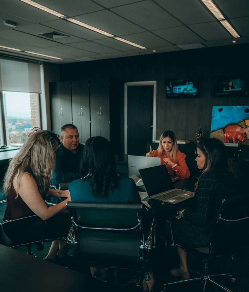 A diverse group of business professionals sit around a conference table with laptops, collaborating in a modern office.