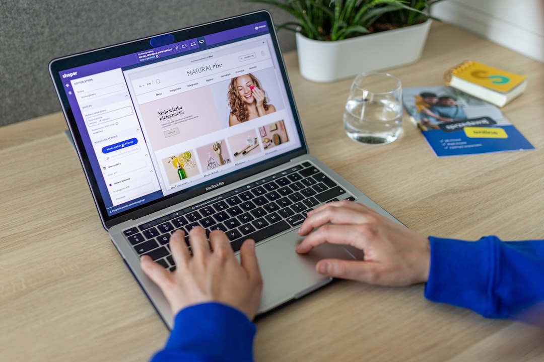 Person typing on a MacBook Pro displaying an online shop with a purple header on a wooden desk.