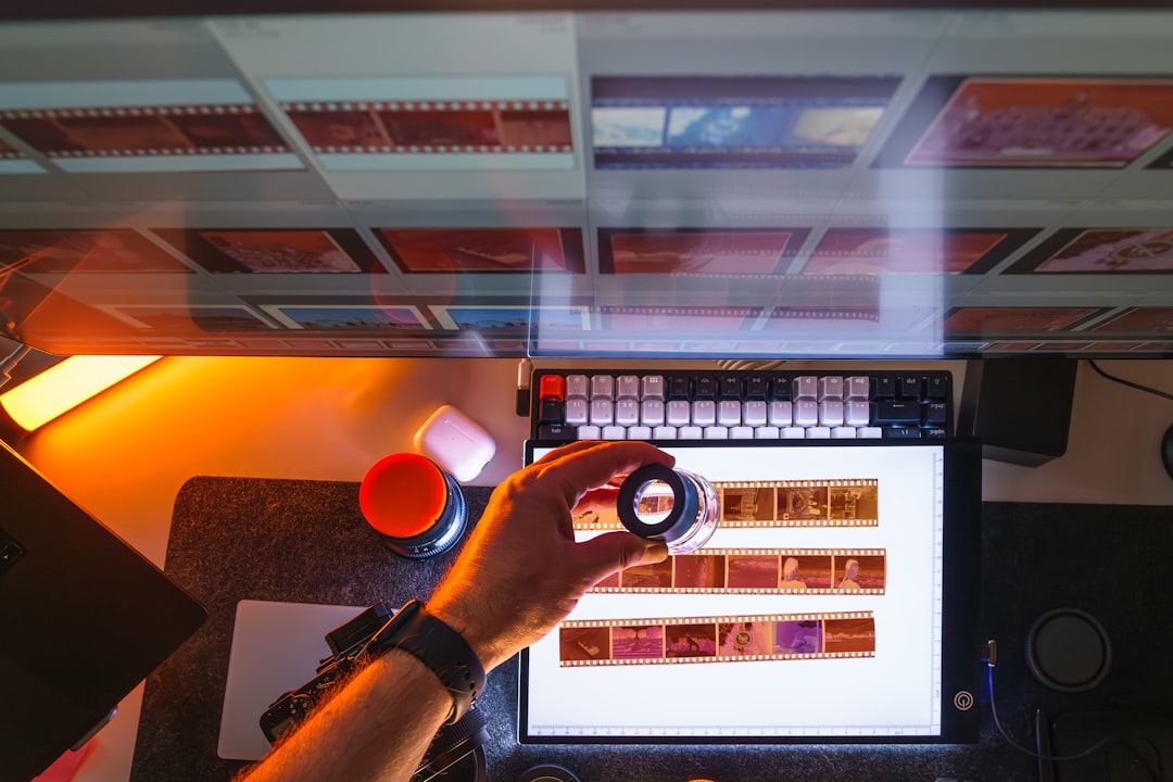 Overhead view of a film-editing setup: a hand holds a lens above a monitor displaying film strips, with camera gear nearby.