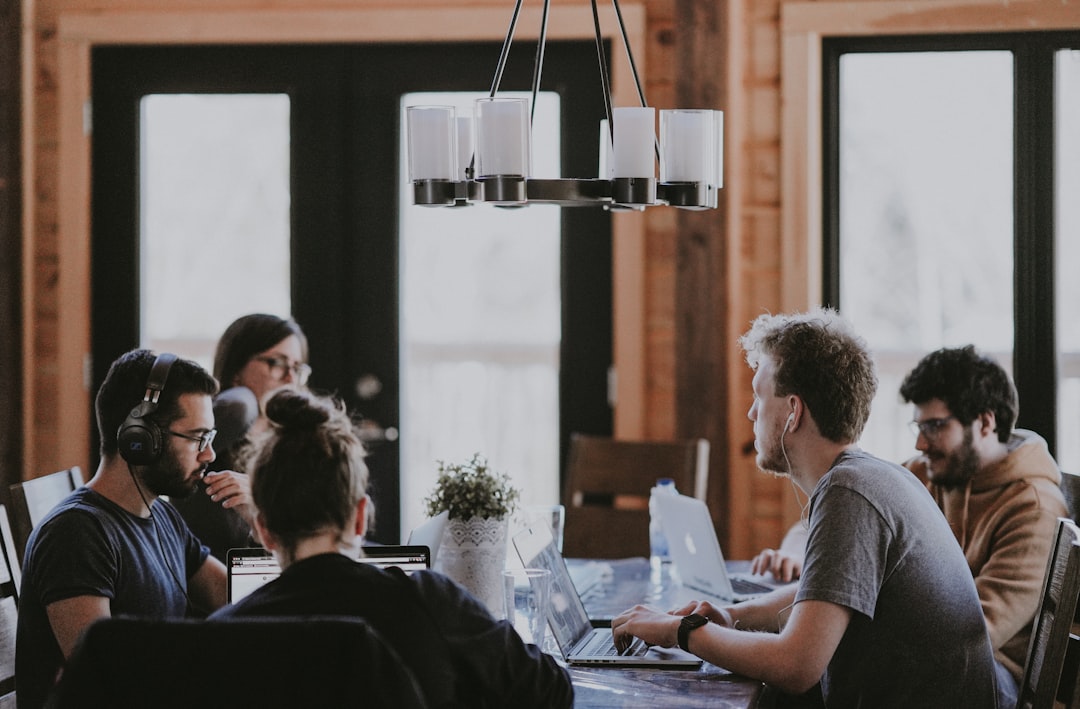 Five people work on laptops around a wooden table in a bright coworking space.