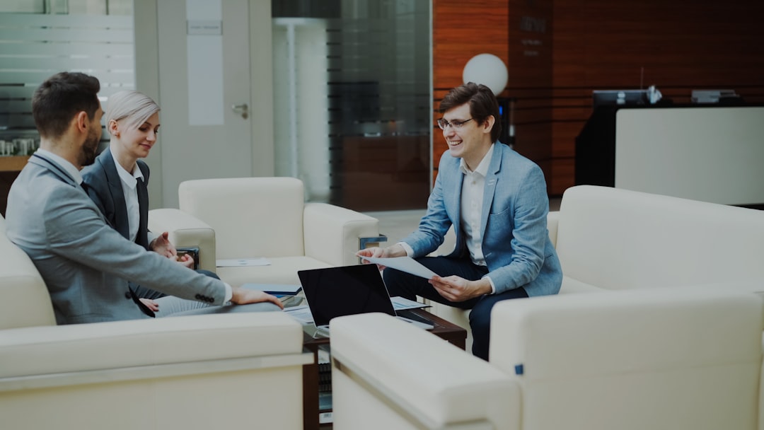 Three colleagues in a modern office sit on white couches, discussing documents with a laptop on the table.
