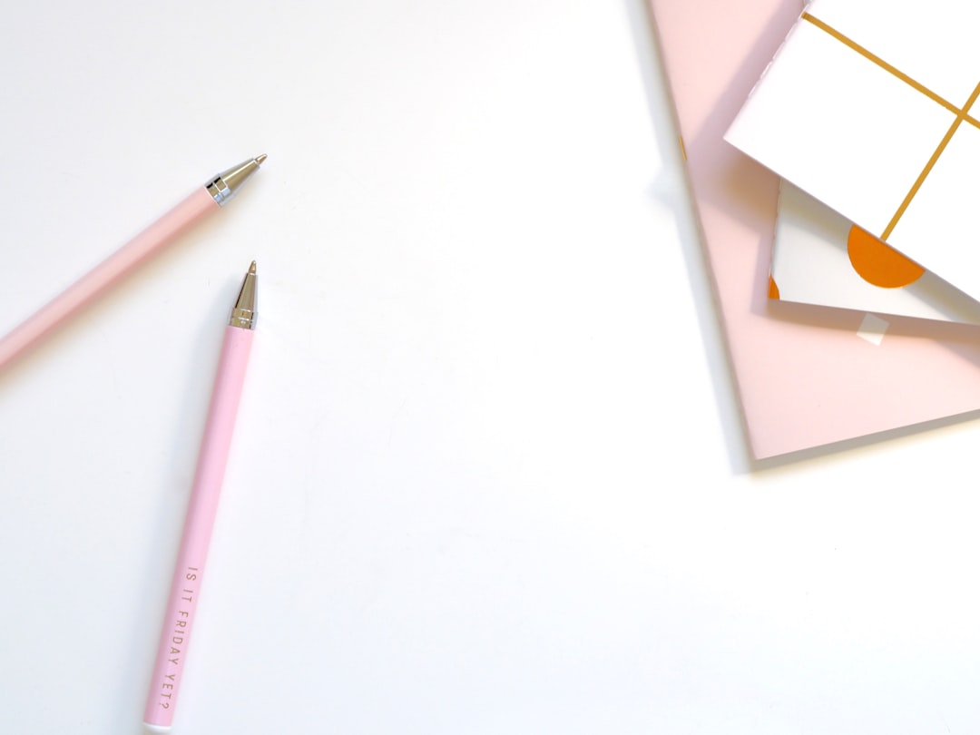 Flat lay of two pink pencils on a white desk with pastel notepads in the top-right corner.