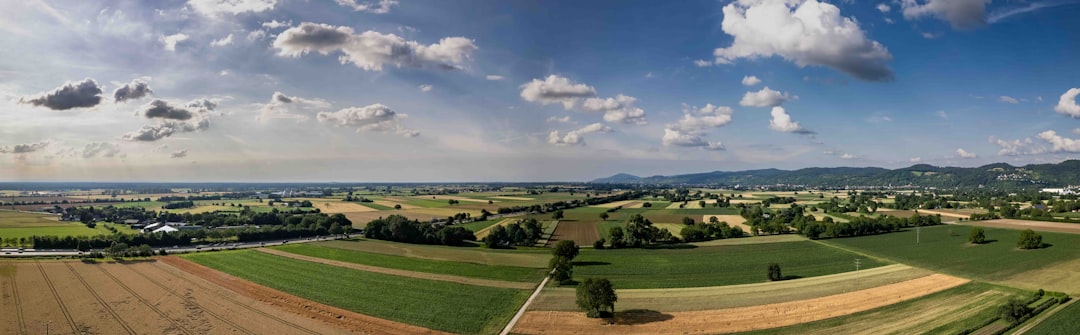 Panoramic view of patchwork farmland with fields, roads, and distant hills under a blue sky with clouds.