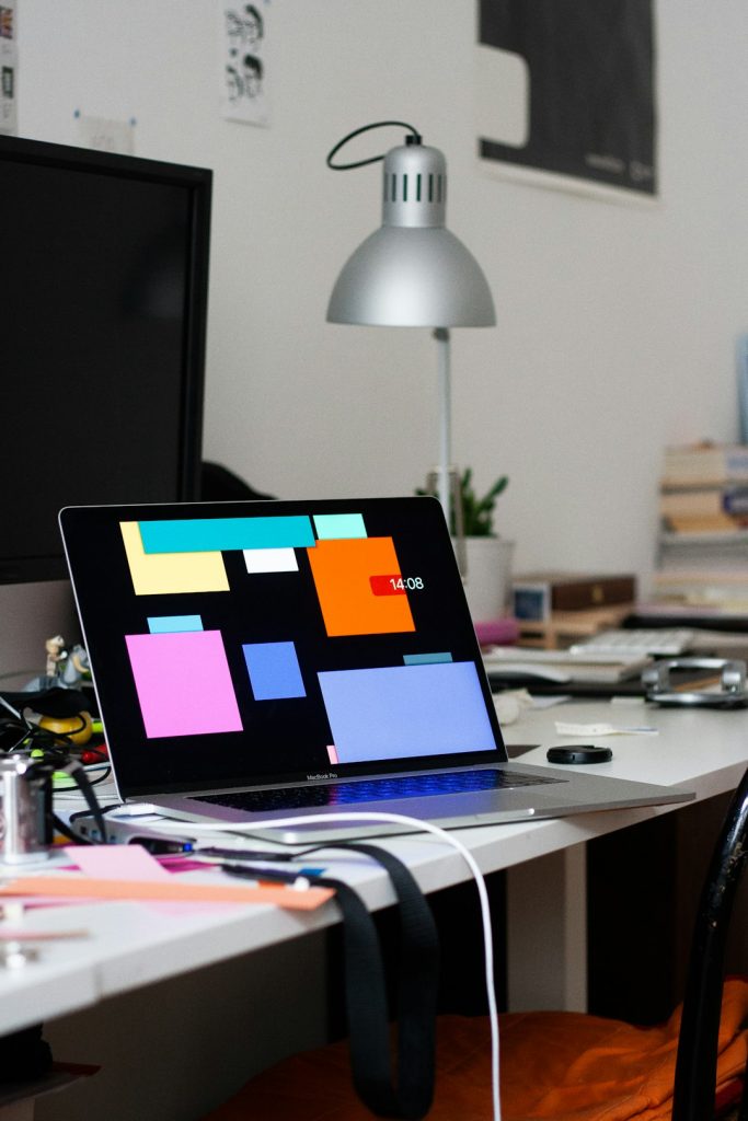 Open MacBook Pro on a cluttered desk, screen filled with colorful Post-it notes and office supplies nearby (lamp in background).