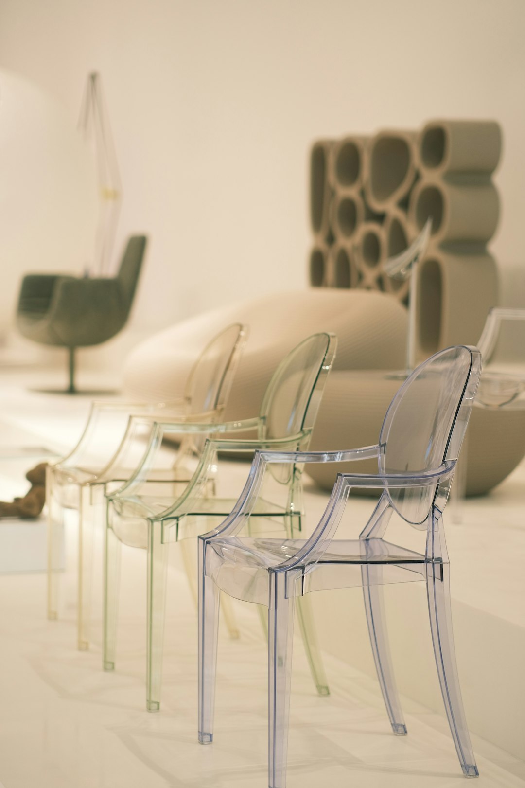 Row of transparent acrylic chairs in a bright minimalist showroom, with an abstract sculptural piece blurred in the background.