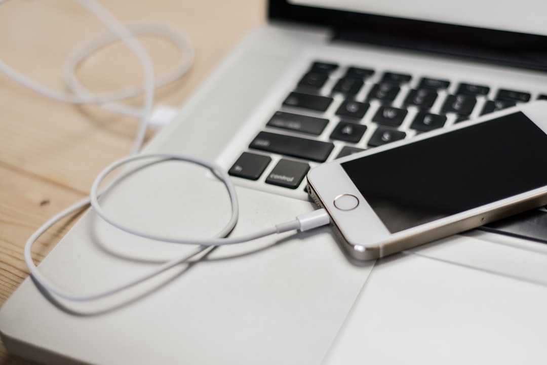 Phone charging from a laptop via USB cable on a wooden desk area near the keyboard.