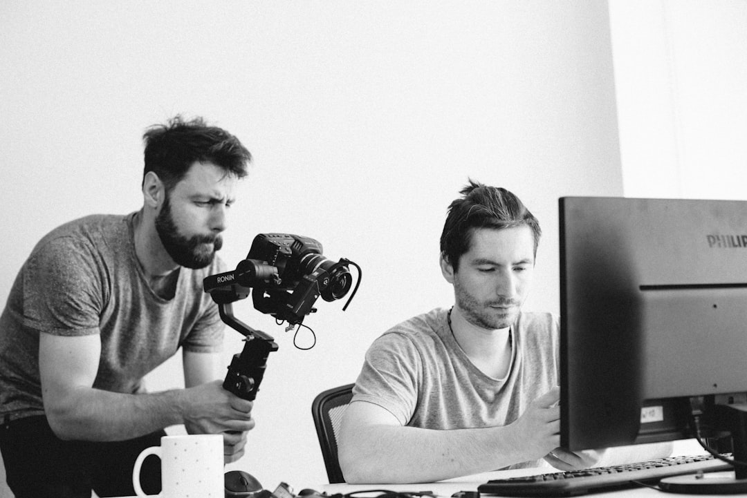 man holding camera pointing at monitor beside sitting man team collaboration video editing remote work - AI Bud