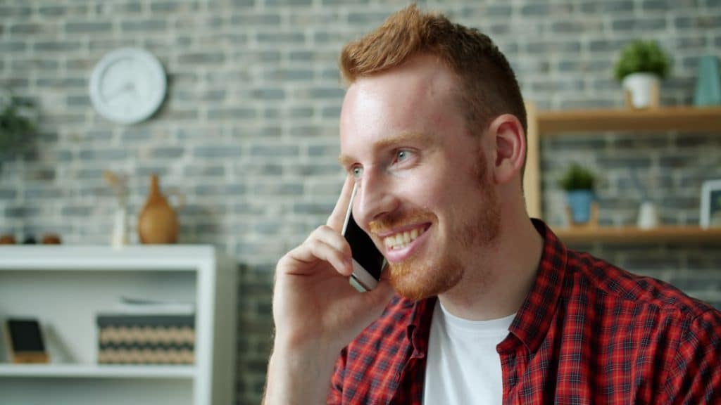 Man in a red plaid shirt smiles while talking on his smartphone in a cozy home office setting with a brick wall backdrop.