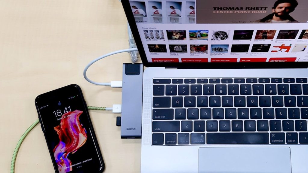 Laptop displaying a video gallery on screen with a phone plugged into a USB hub beside it on a light wooden desk.