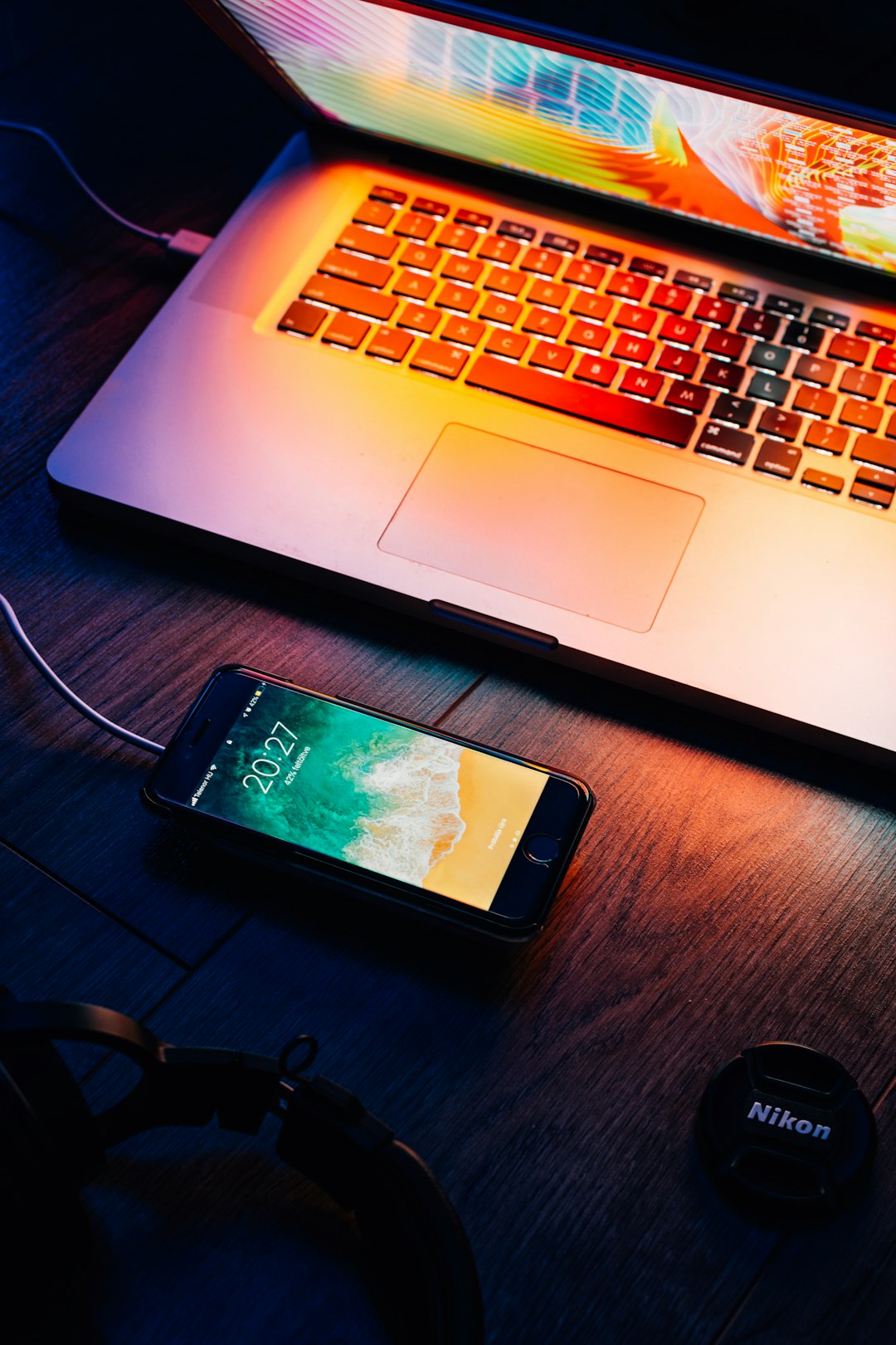 Laptop with multicolored neon lighting on keyboard, charging iPhone on a wooden desk, Nikon lens cap nearby.