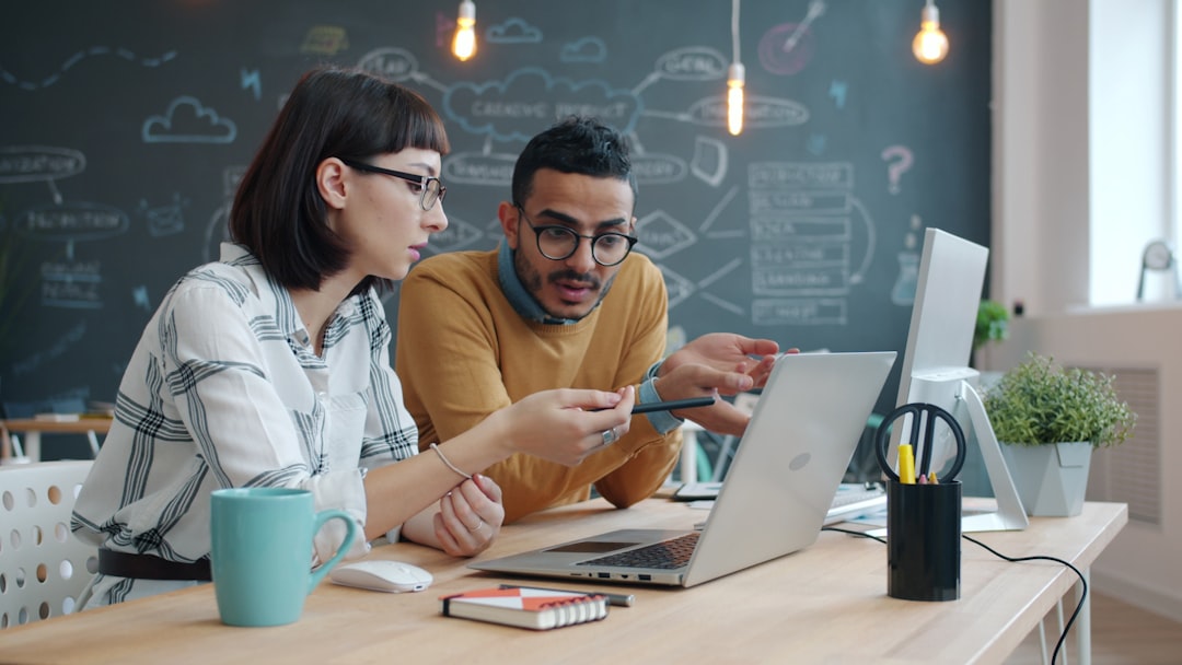 Two colleagues collaborate at a desk with a laptop, notebook, and pens, one pointing at the screen as they discuss work in front of a chalkboard filled with diagrams.