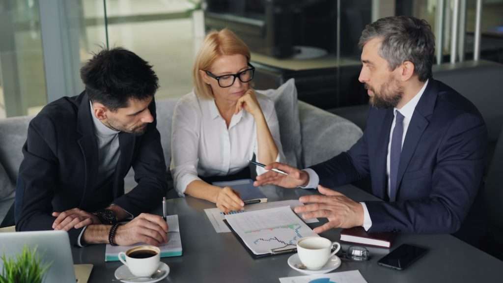 Three professionals in a meeting, reviewing printed charts and notes on a conference table.