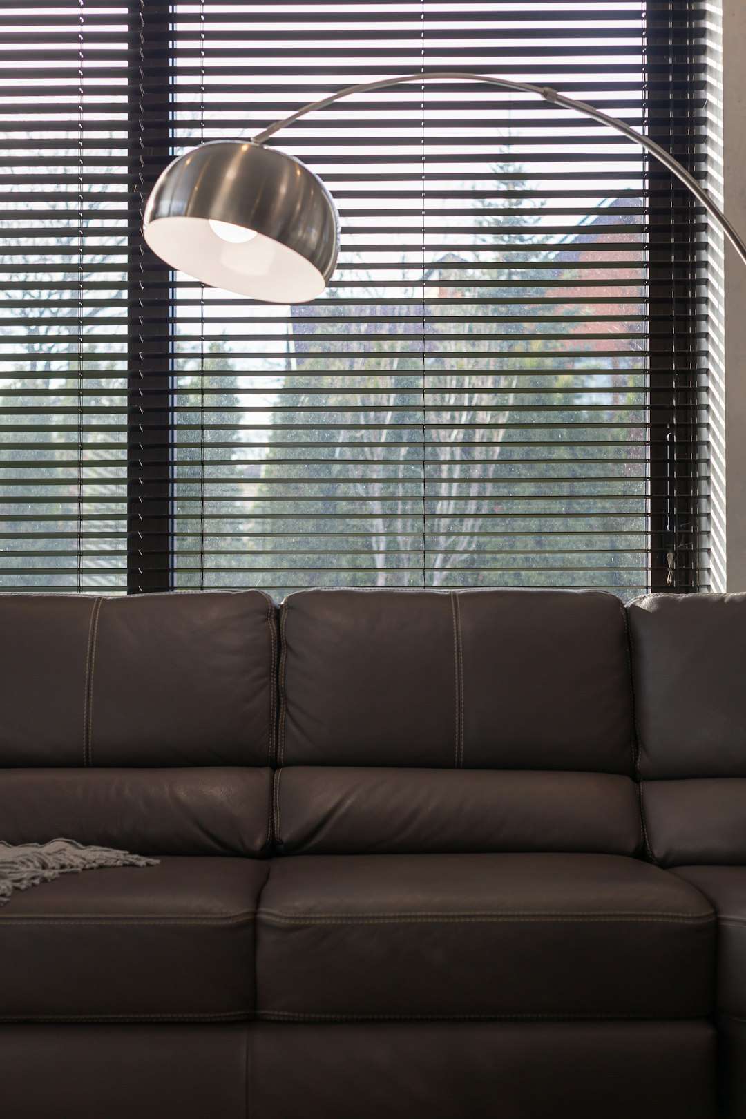 Dark brown leather sofa in front of a large window with horizontal blinds; a curved arc floor lamp arches overhead.