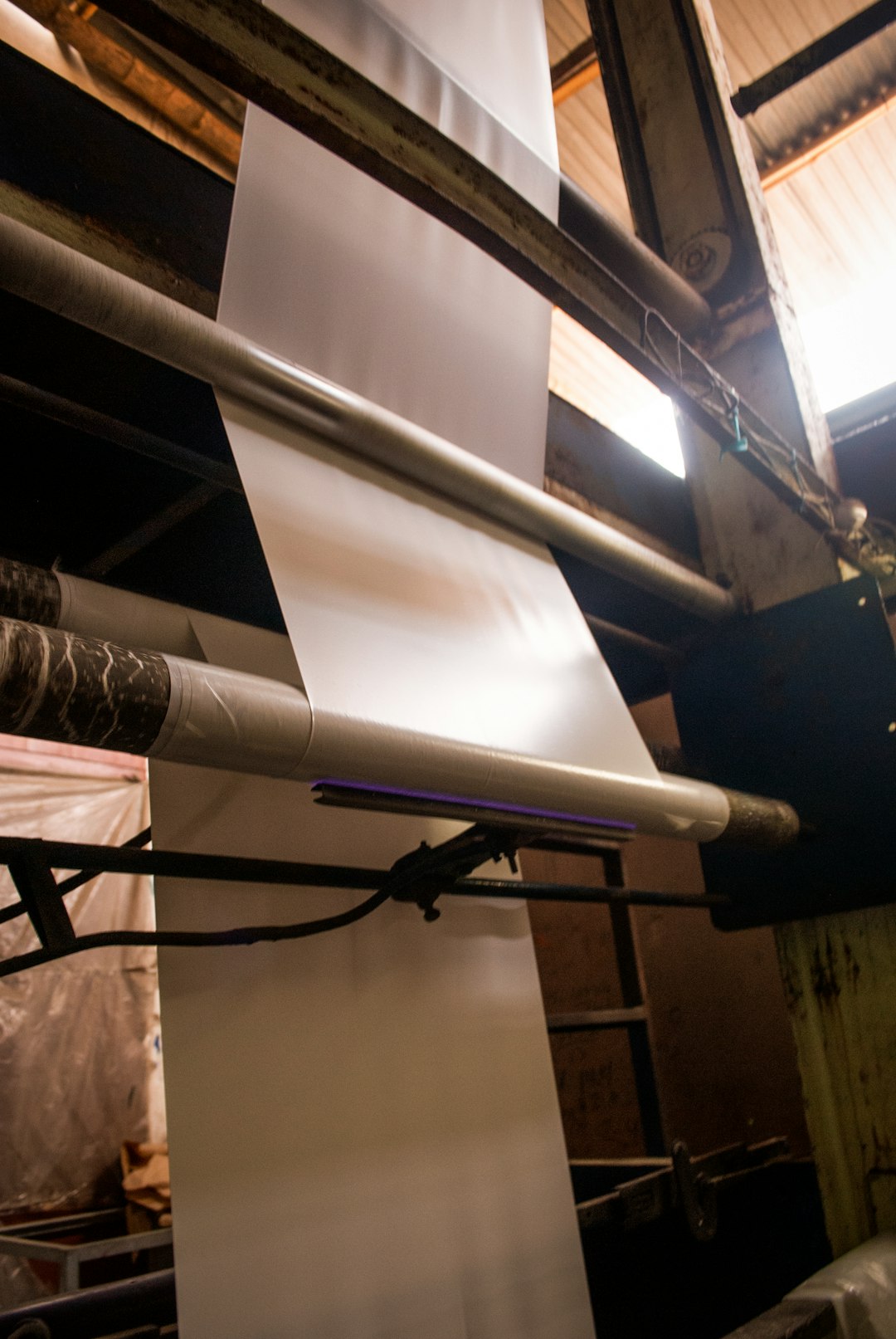 White sheet being fed over a cylindrical roller in an industrial production area, with exposed beams and pipes in the background.