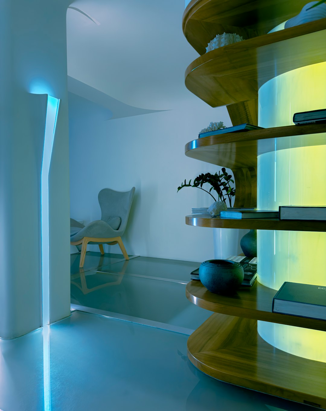 Modern interior with curved wooden shelves emitting blue-green glow, books and potted plant on display, contemporary chair in background.