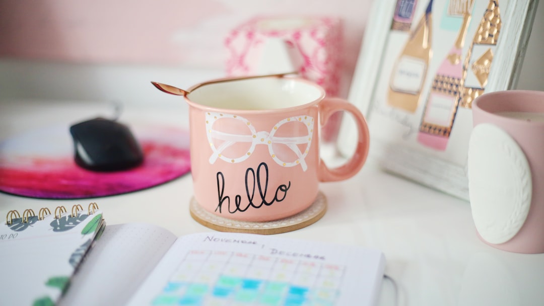 Pink mug with white glasses illustration and 'hello' text on a desk, with notebook, mouse, and pastel decor nearby.
