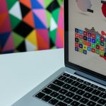 Laptop on a white desk with a bright geometric mural behind; screen shows a colorful grid of social icons.