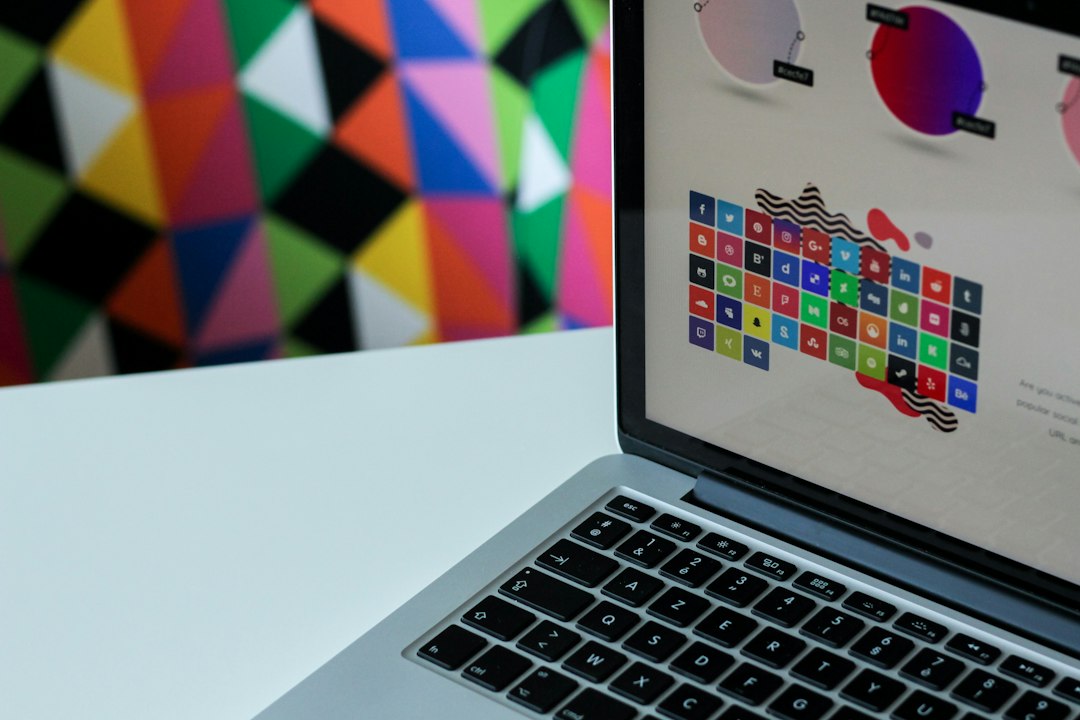 Laptop on a white desk with a bright geometric mural behind; screen shows a colorful grid of social icons.