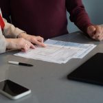 Two people review printed documents at a table, with a pen, smartphone, and tablet nearby.