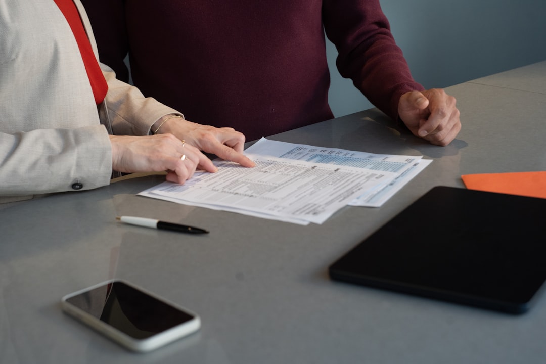 Two people review printed documents at a table, with a pen, smartphone, and tablet nearby.