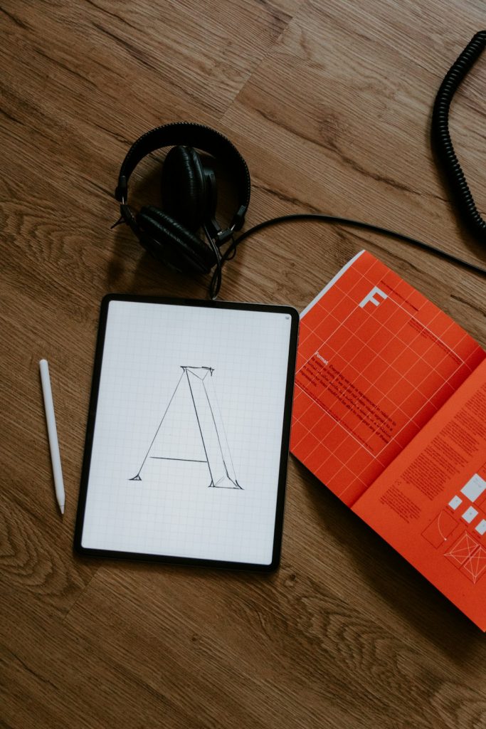Workspace with a tablet showing a sketch of an A-frame structure, a white stylus nearby, and orange books on a wooden floor