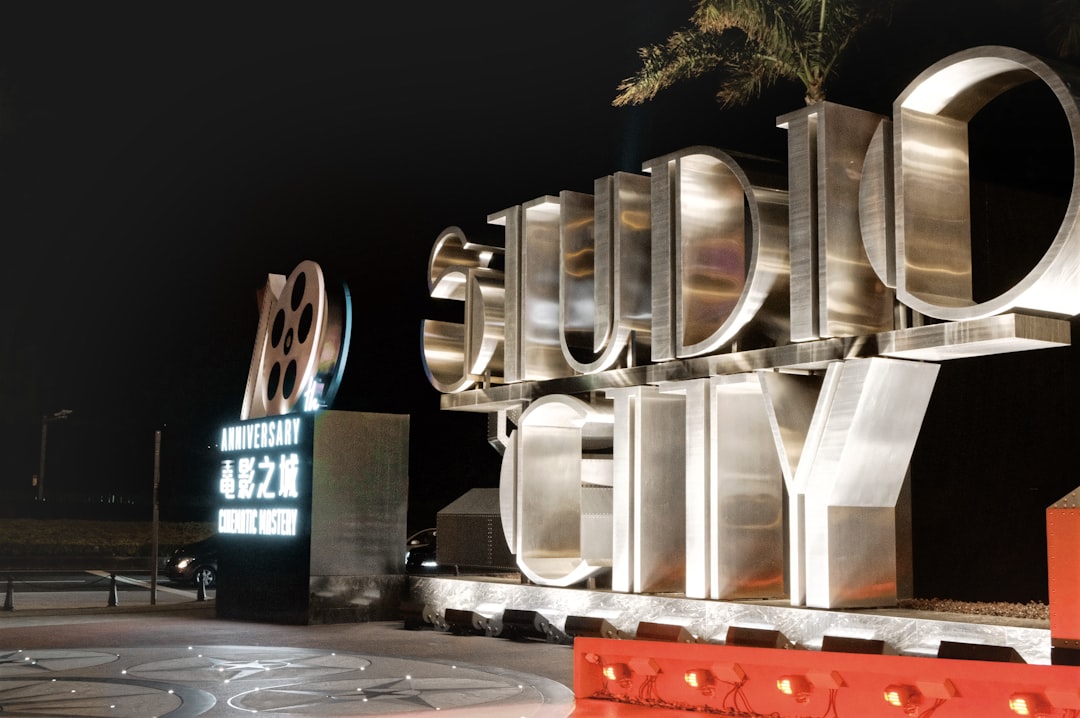 Nighttime view of a large metallic lettering sculpture forming a word, with a film reel emblem to the left and a glowing sign beside it.