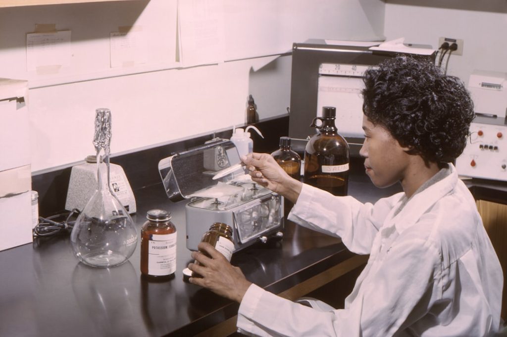 Lab technician in a white coat handling a bottle on a benchtop with glassware, jars, and a scale in a chemistry lab.