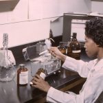 Lab technician in a white coat handling a bottle on a benchtop with glassware, jars, and a scale in a chemistry lab.
