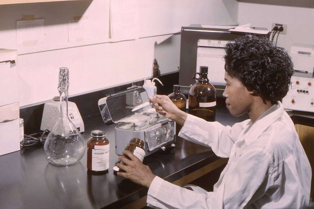 Lab technician in a white coat handling a bottle on a benchtop with glassware, jars, and a scale in a chemistry lab.