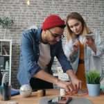 Two coworkers collaborate on a small craft project at a wooden table in a brick-walled open office environment.