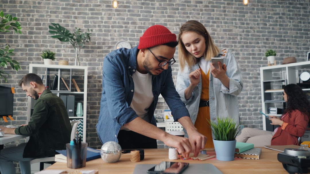 Two coworkers collaborate on a small craft project at a wooden table in a brick-walled open office environment.