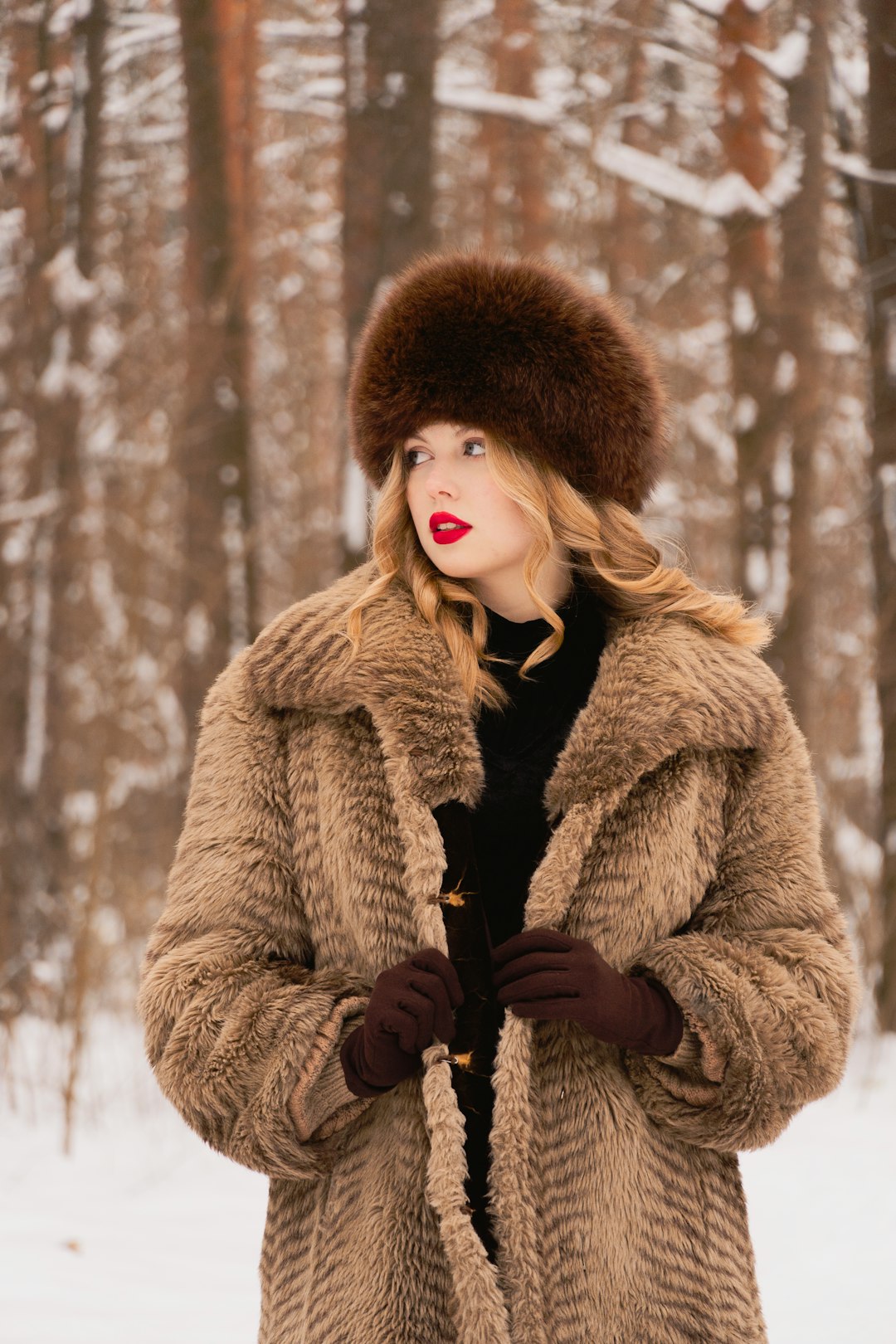 Woman in a thick brown fur coat and matching hat standing in a snowy forest, looking off to the side with red lipstick.