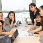 Seven young professionals around a conference table collaborate, reviewing a colorful poster with charts as laptops and notebooks are open.