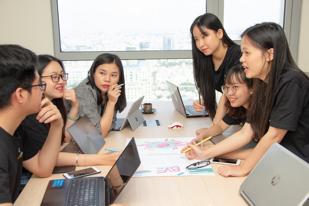 Seven young professionals around a conference table collaborate, reviewing a colorful poster with charts as laptops and notebooks are open.
