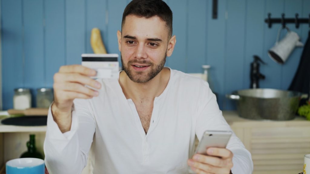 Man in a white shirt holds a credit card up while looking at his smartphone in a blue-kitchen setting, suggesting online shopping or payment.